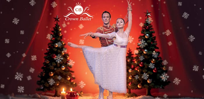 Ein Balletttänzer in rotem Kostüm hält eine Tänzerin in weißem Kleid in Pose, umgeben von weihnachtlich geschmückten Tannenbäumen und Kerzen, vor rotem Hintergrund mit Schneeflocken.