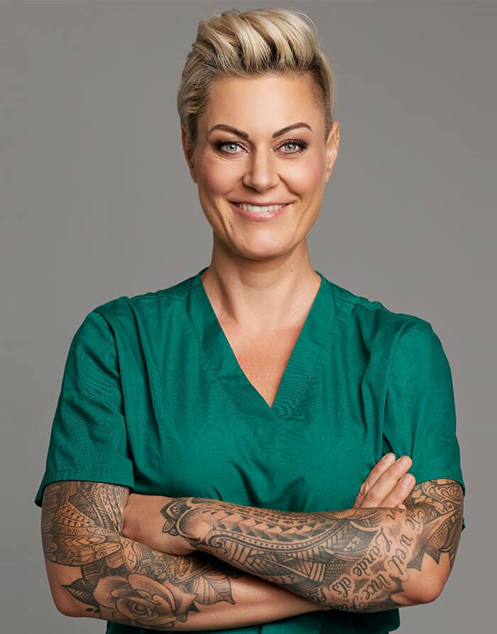 Eine blonde Frau mit verschränkten Tattoowierten Armen in einem grünem Ärztekittel ist zu sehen.
