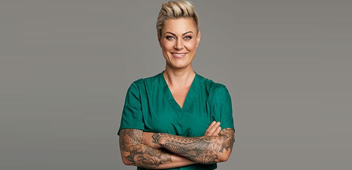 Eine blonde Frau mit verschränkten Tattoowierten Armen in einem grünem Ärztekittel ist zu sehen.