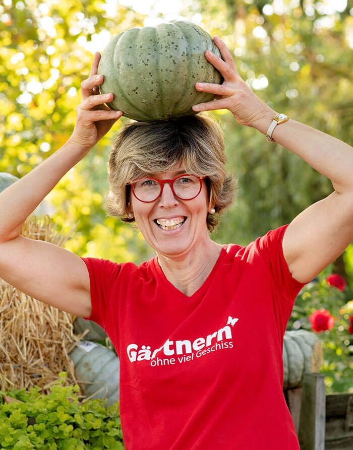 Eine Frau mit rotem T-Shirt und roter Brille hält einen grünen Kürbis lachend auf dem Kopf, im Hintergrund Strohballen und Gartenpflanzen.