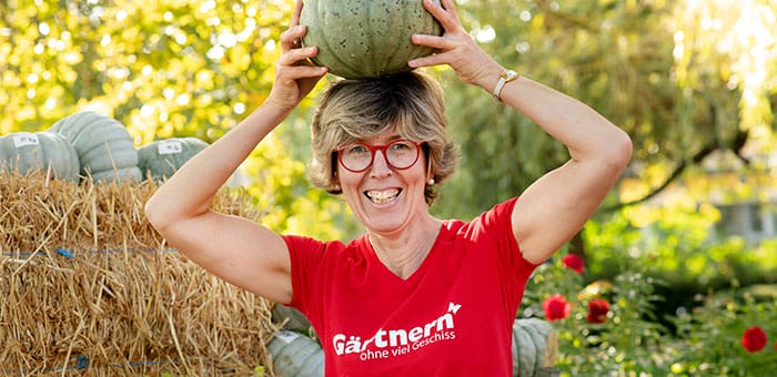 Eine Frau mit rotem T-Shirt und roter Brille hält einen grünen Kürbis lachend auf dem Kopf, im Hintergrund Strohballen und Gartenpflanzen.