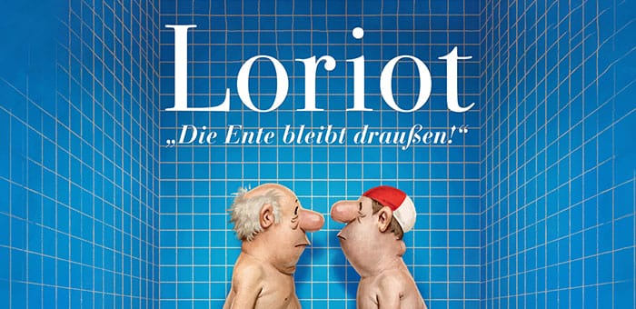 Man sieht ein Blau gekacheltes Badezimmer, in der Mitte hinter dem schwarz weißen Schachbrett Boden ist die Badewanne in der sich zwei gezeichnete Männer Nackt gegenüber stehen und anschauen, der rechte Mann trägt eine weiß rote Badehaube. Vor der Wanne steht ein Hocker auf dem eine Braune Jacke liegt und darauf eine Badeente. Auf dem Teppich vor der Wanne liegen zwei Hausschuhe.