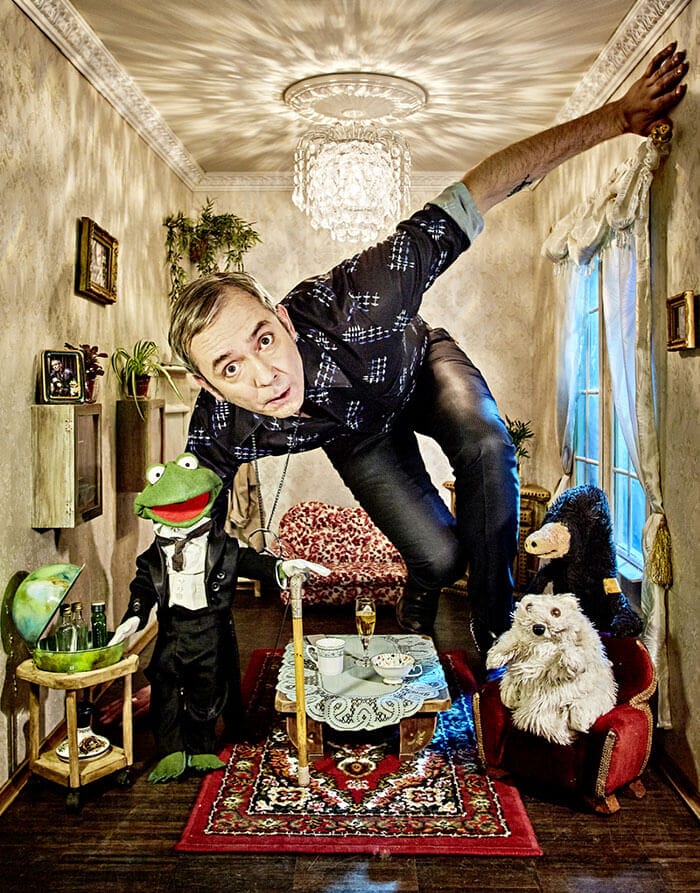 Ein Mann beugt sich in ein kleines Wohnzimmer voller Puppen, darunter ein Frosch im Smoking, ein weißer Hund und ein schwarzer Bär, die an einem gedeckten Tisch sitzen.