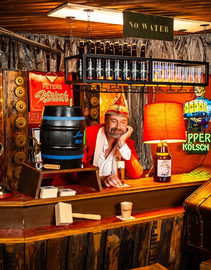 Ein Mann in rotem Jackett und Karnevalsmütze sitzt lächelnd an einer Theke mit Bierfass, Flaschen und Leuchtschildern im Hintergrund.