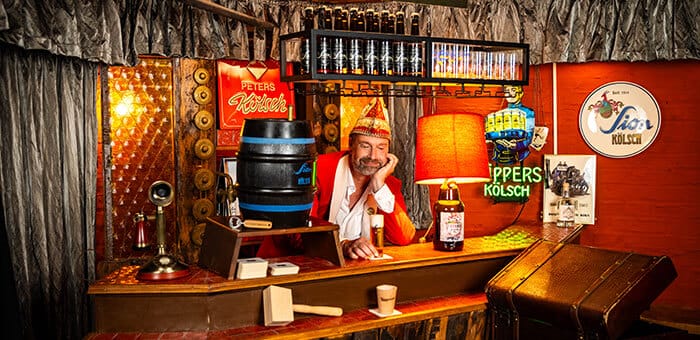 Ein Mann in rotem Jackett und Karnevalsmütze sitzt lächelnd an einer Theke mit Bierfass, Flaschen und Leuchtschildern im Hintergrund.
